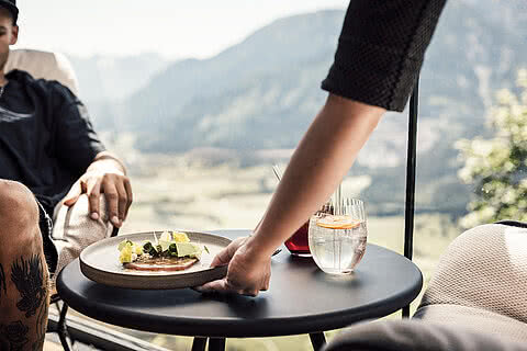 Servieren eines Gerichtes auf der Panoramaterrasse des Restaurants 1250 mit Bergblick, daneben erfrischende Getränke und ein Gast im Hintergrund