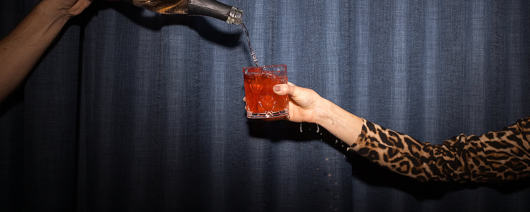 Rosé Prosecco wird in ein Glas gegossen, während eine Hand mit einem animalischen Muster das Glas hält vor einem blauen Vorhang